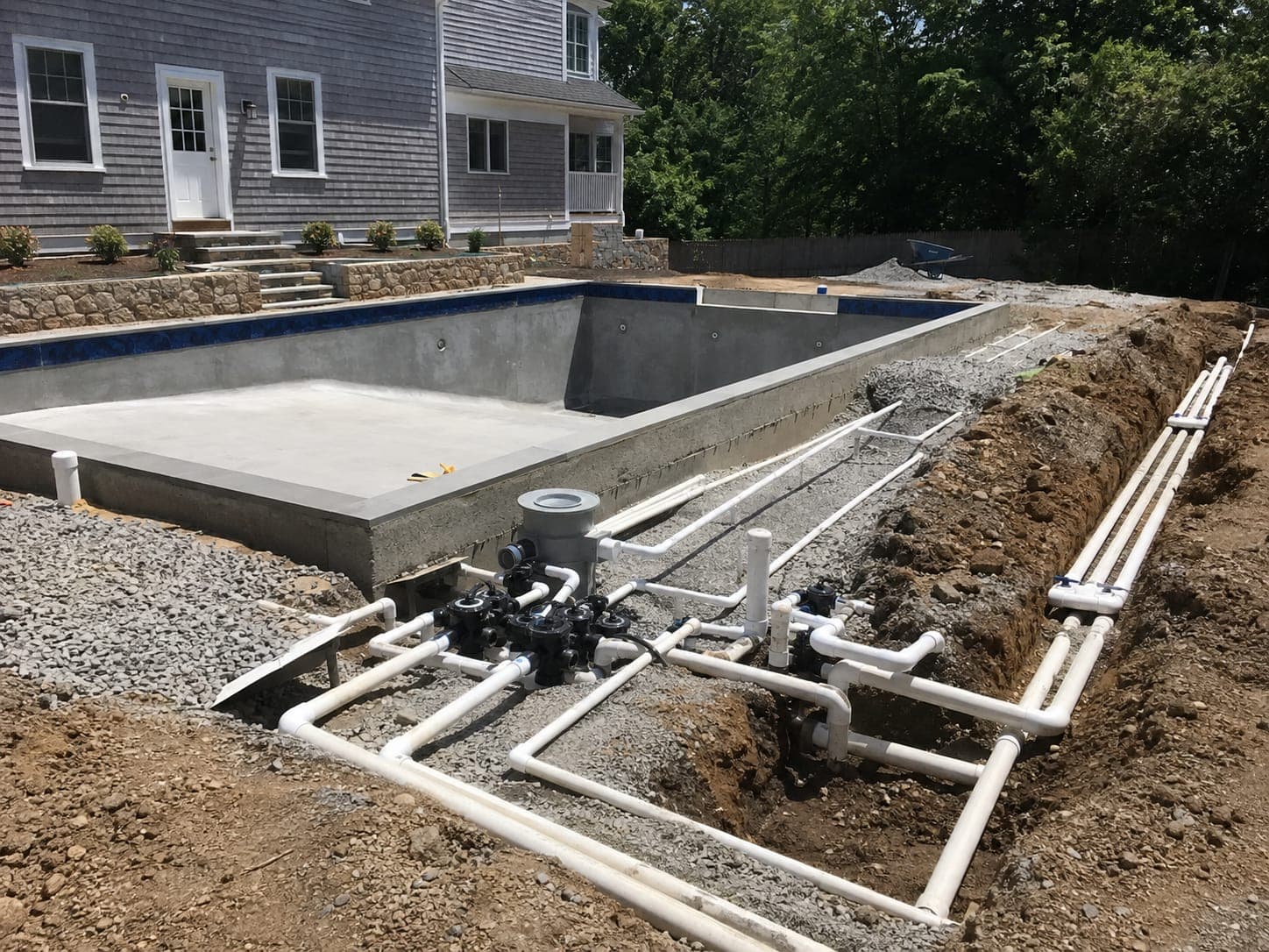 Newly installed white PVC pool plumbing lines and valve manifold in an open trench beside a pool under construction
