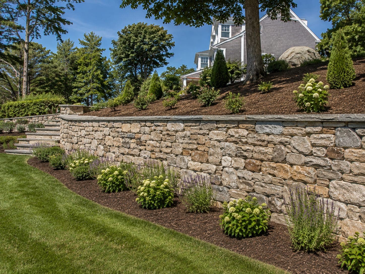 Curved segmental block retaining wall with a bluestone cap holding a planted slope in a Cape Cod yard