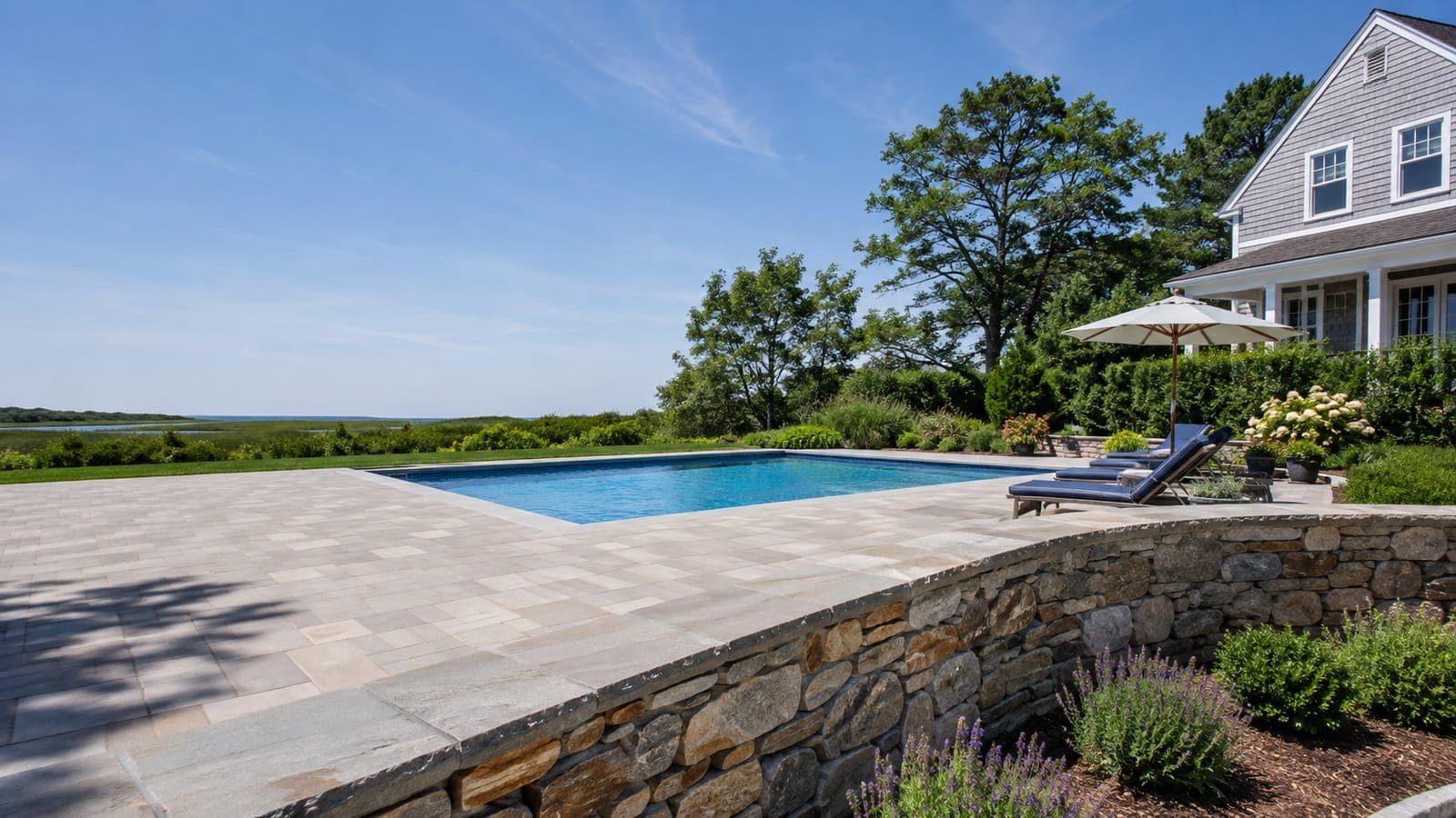 Paver pool patio with a capped stone retaining wall behind a cedar-shingled Cape Cod home